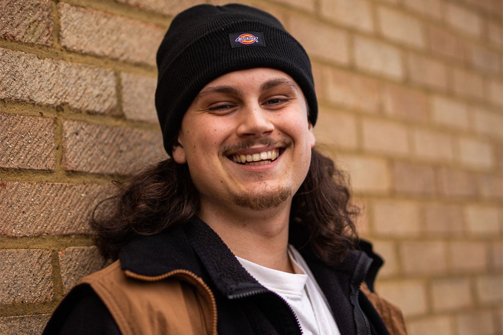 A press shot of Gabe Steel [IZUK] smiling in front of a brick wall