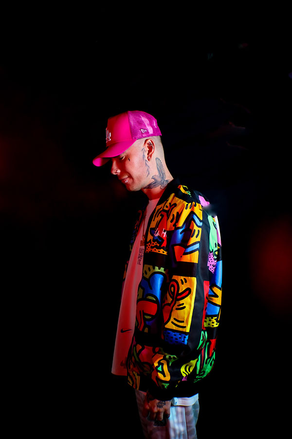 A portrait of Ben Cross (Crossy) wearing a colourful jacket, pink baseball hat with a dark backdrop behind him