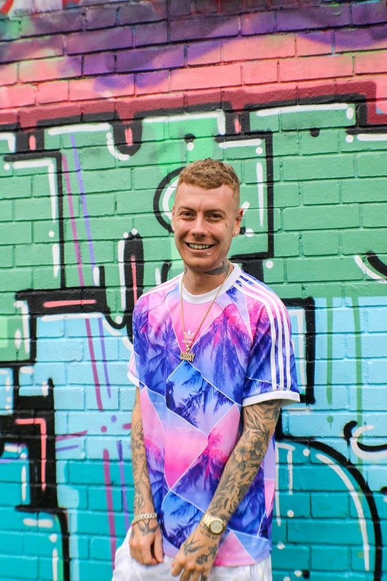 A portrait of Ben Cross (Crossy) smiling into camera with a graffiti'd wall in the background