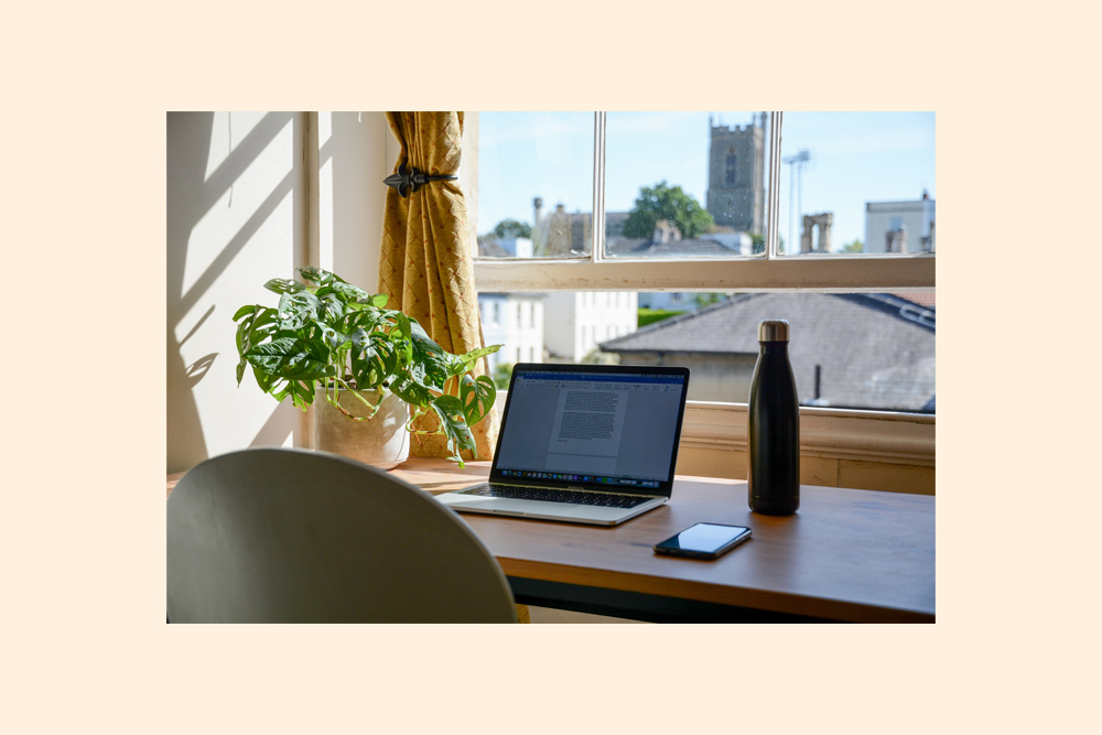 A picture of a working desk, with a monstera plant, laptop, mobile phone and water bottle. Outside the window is a village and church